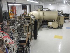 A tandem particle accelerator. A lab with a lot wiring and a tan, cylindrical accelerator. 