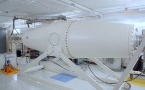 The body of a tandem particle accelerator. It is a long, white object shaped like a cylinder that is symmetrically tapered at each end. 