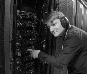 Will wearing Ear Defenders, pointing at the back of a rack of SCARF compute nodes.