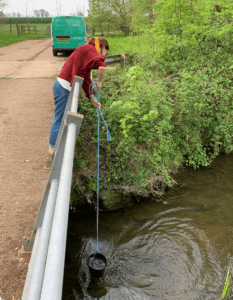 Katie is taking water from a river, from a bridge above, using a bucket attached to a rope