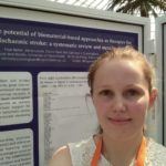 Selfie of me standing in front of a white and purple poster. There's a palm tree in the background.