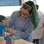 Rebekah is in the lab in a blue lab coat holding a pipette.