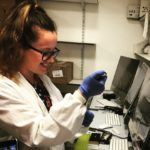 Rebekah is stood in front of computers which are attached to sequencing devices, and she is holding a pipette and a tube preparing a sequencing library.