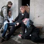 Four people in costume. One is a soldier threatening the others. One is in a lab coat screaming. One is slumped on another's knee, apparently unconscious.