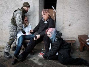 Four people in costume. One is a soldier threatening the others. One is in a lab coat screaming. One is slumped on another's knee, apparently unconscious.