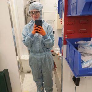 A woman wearing a cleanroom suit, goggles and bright orange nitrile gloves