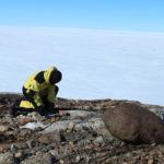 This is a scientist collecting a rock sample in Antarctica. They are kneeling on a rocky outcrop with the ice sheet behind them.