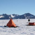 An orange tent in front of mountains in Antarctica. The tent is on the ice sheet. This is where people live when they are in the field.