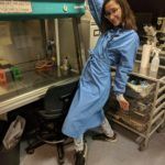 A women with brown hair in a blue lab coat, posing with open arms in front of a tissue culture hood