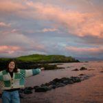 A pink sunset over the sea, a headland and a some rocks in the sea with me with my arms outstretched wearing a patchwork cardigan with squares of different shades of green, cream and beige.