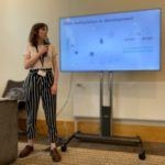 Katie standing with a microphone in front of a white screen talking about her research.
