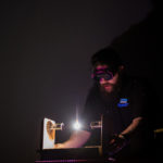Dan is wearing welding gollgles as he produces a very bright white light using an electrical arc. The rest of the room is dark.