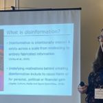 Laura standing next to a projector screen showing a slide called 'What is disinformation'. The slide is illustrated with a picture of a laptop