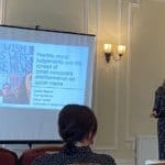 Laura stands next to a projector screen showing the title slide of her presentation 'Flexible moral judgements and the spread of belief-consistent disinformation'. The slide features a picture of a man holding up a handwritten sign saying 'I wish this were fake news'