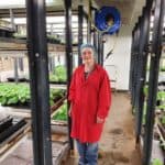 Caroline in front of shelves containing trays of small green plants in pots