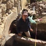 White woman in early 20s standing in a trench behind a half wall made of brick, using a measuring tape and smiling at the camera